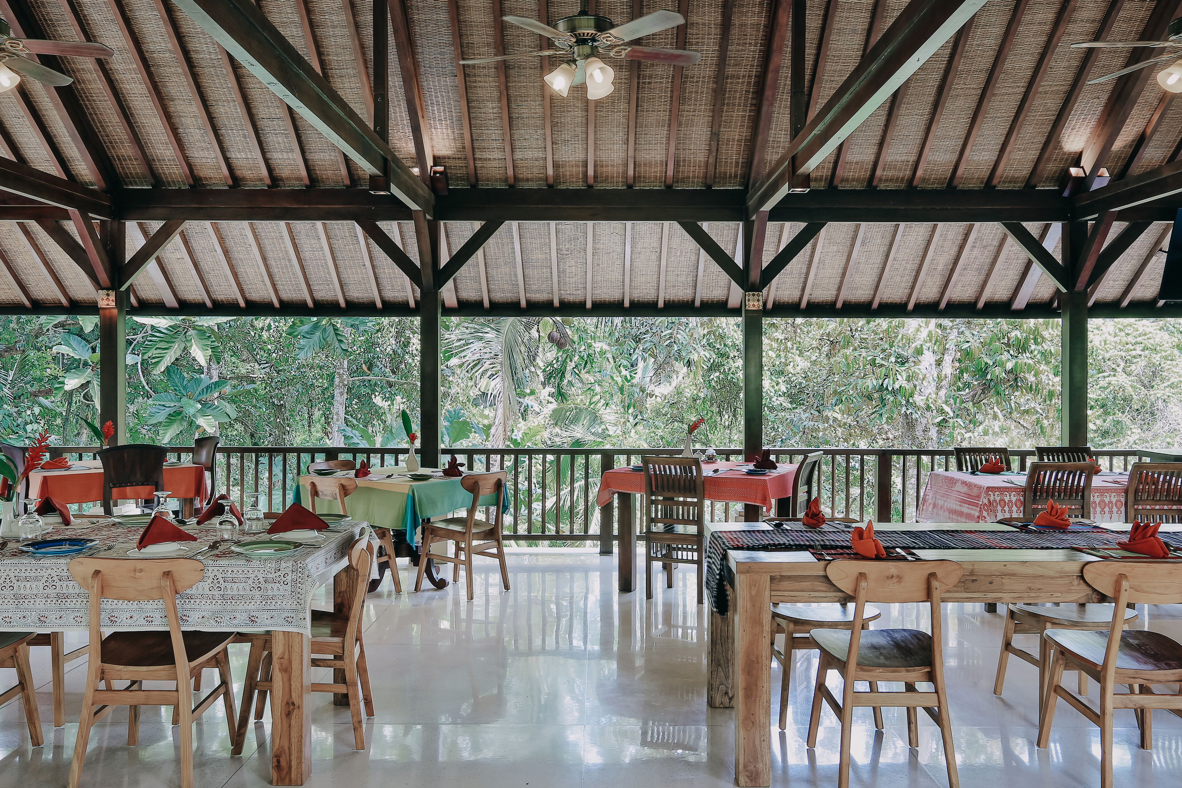 An open-air restaurant with wooden tables and chairs, a ceiling fan, and a view of lush greenery.