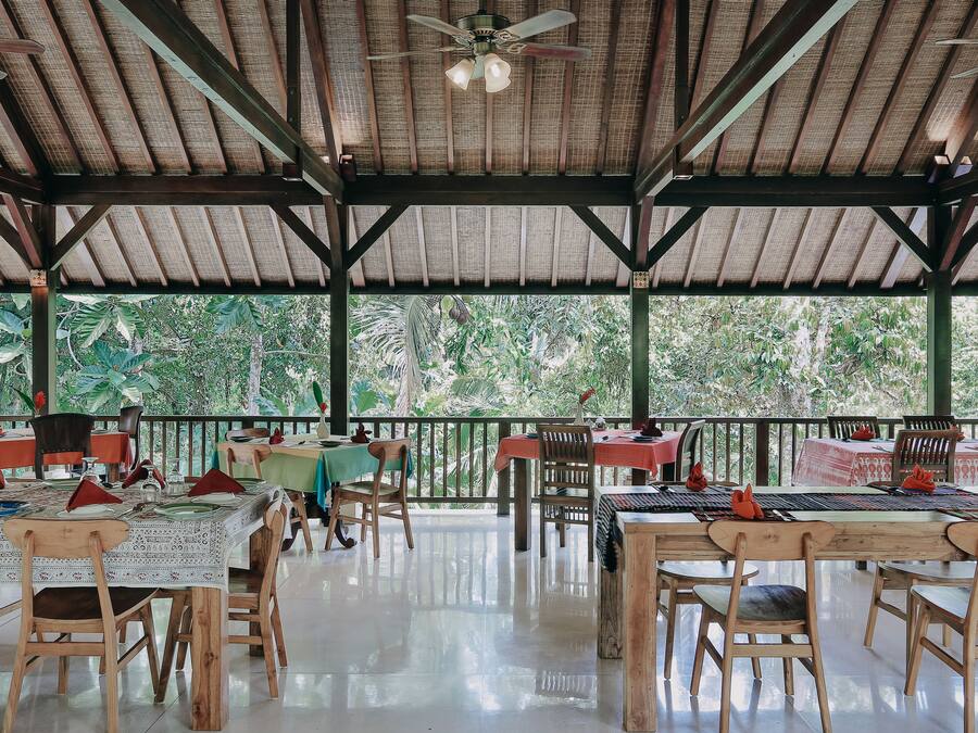 An open-air restaurant with wooden tables and chairs, a ceiling fan, and a view of lush greenery.