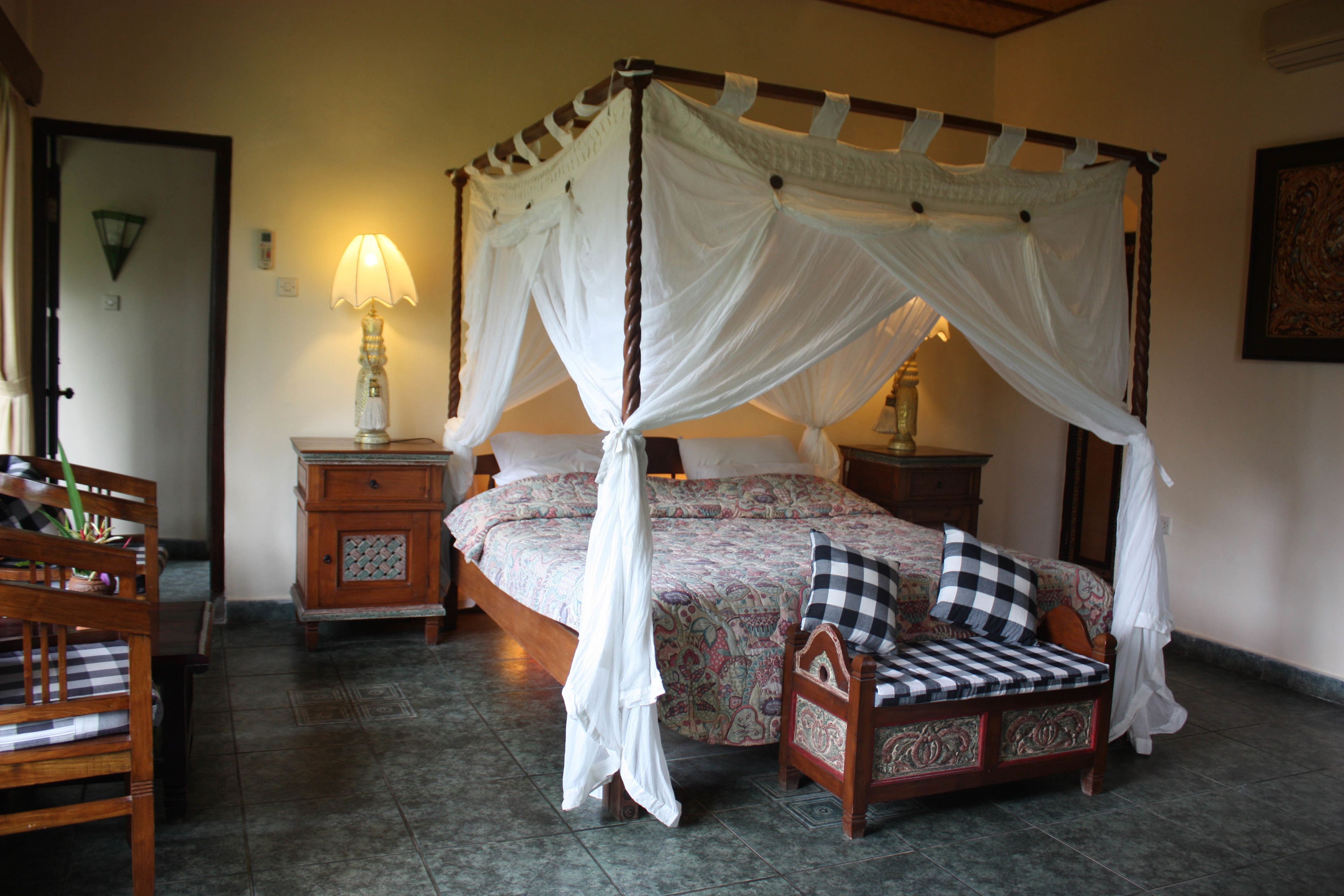 A four-poster canopy bed with a checkered pillow, a nightstand with a lamp, and a wooden chair in a room with tiled flooring.
