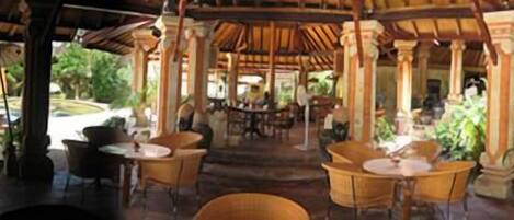 An outdoor covered dining area with tables and chairs, a pool in the background, and tropical plants.