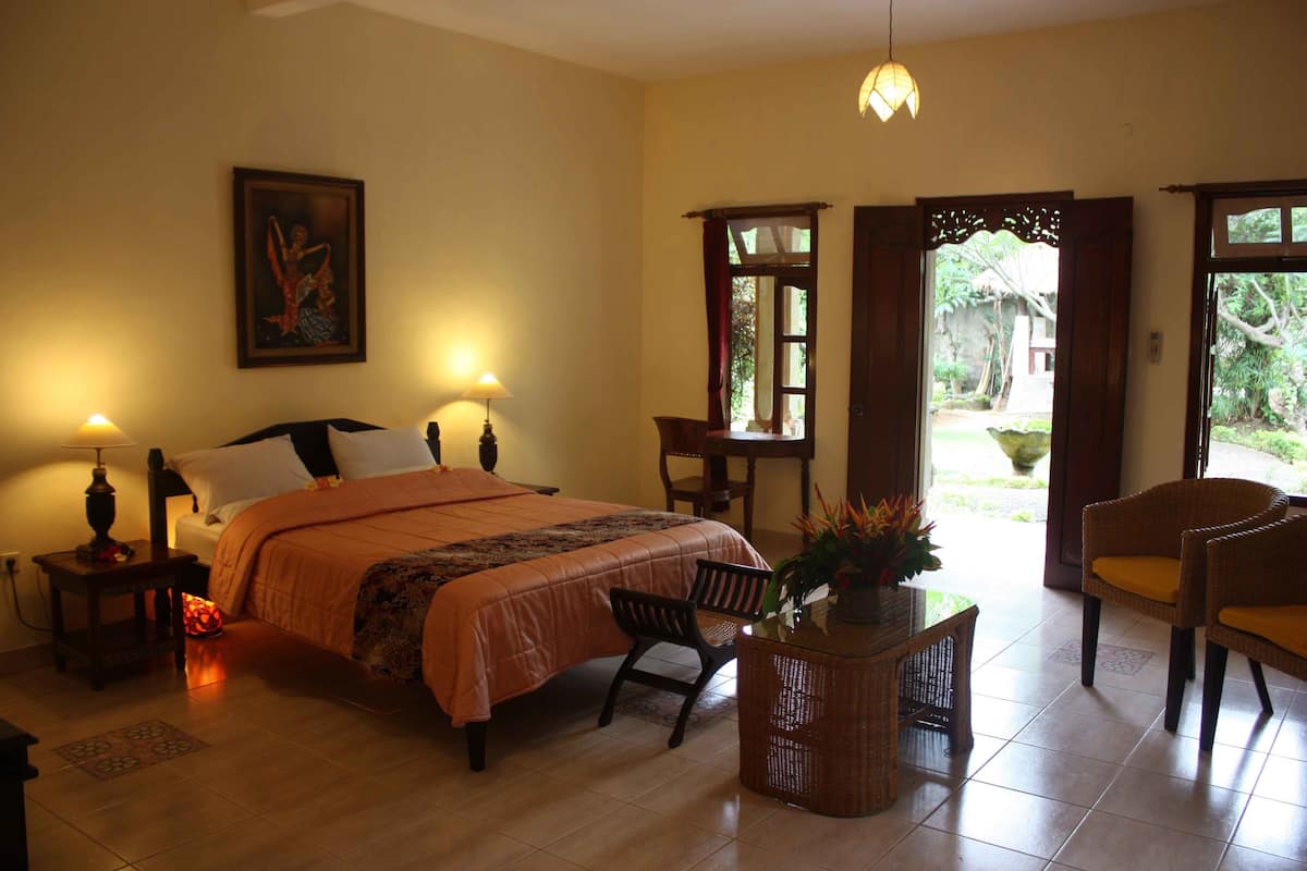 Une chambre à coucher comprenant un lit, des tables de chevet, une commode, un fauteuil en osier, une petite table avec un vase et une porte donnant sur un espace extérieur.