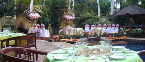 An outdoor dining area with tables set for a meal, surrounded by tropical vegetation and a pool.