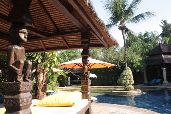 A pool area with a thatched roof structure, a statue, and a well-maintained garden with a palm tree.
