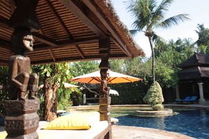 A pool area with a thatched roof structure, a statue, and a well-maintained garden with a palm tree.