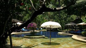 Zona de piscina con una fuente central, rodeada de sillas de descanso y sombrillas, ubicada en un jardín exuberante con vegetación densa.