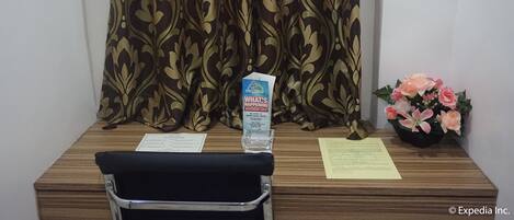 A wooden desk with a black chair, a water bottle, a welcome brochure, and a small flower arrangement.
