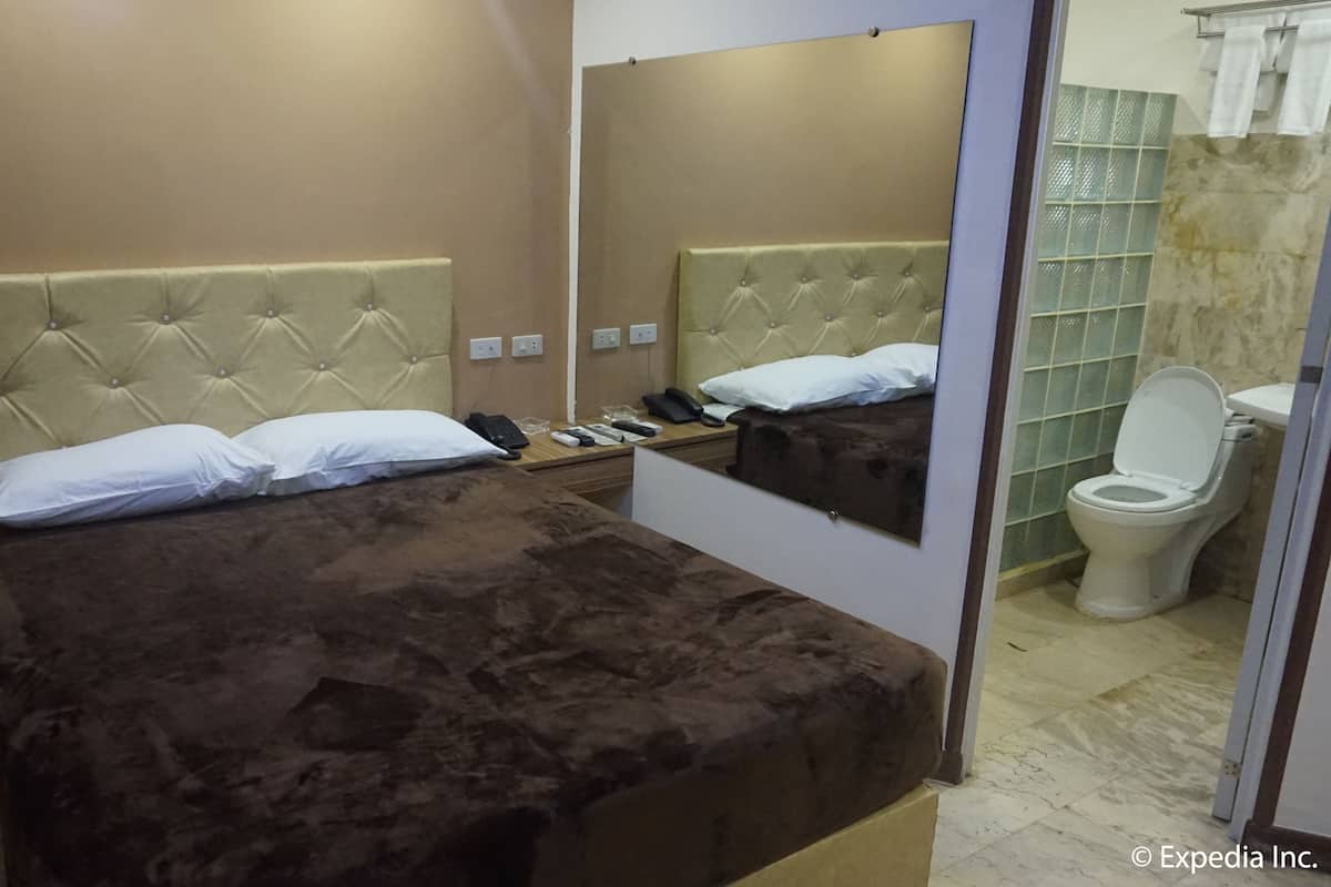 Una habitación de hotel con una cama, una mesita de noche con teléfono y controles remotos, un espejo y un baño con inodoro y ducha.
