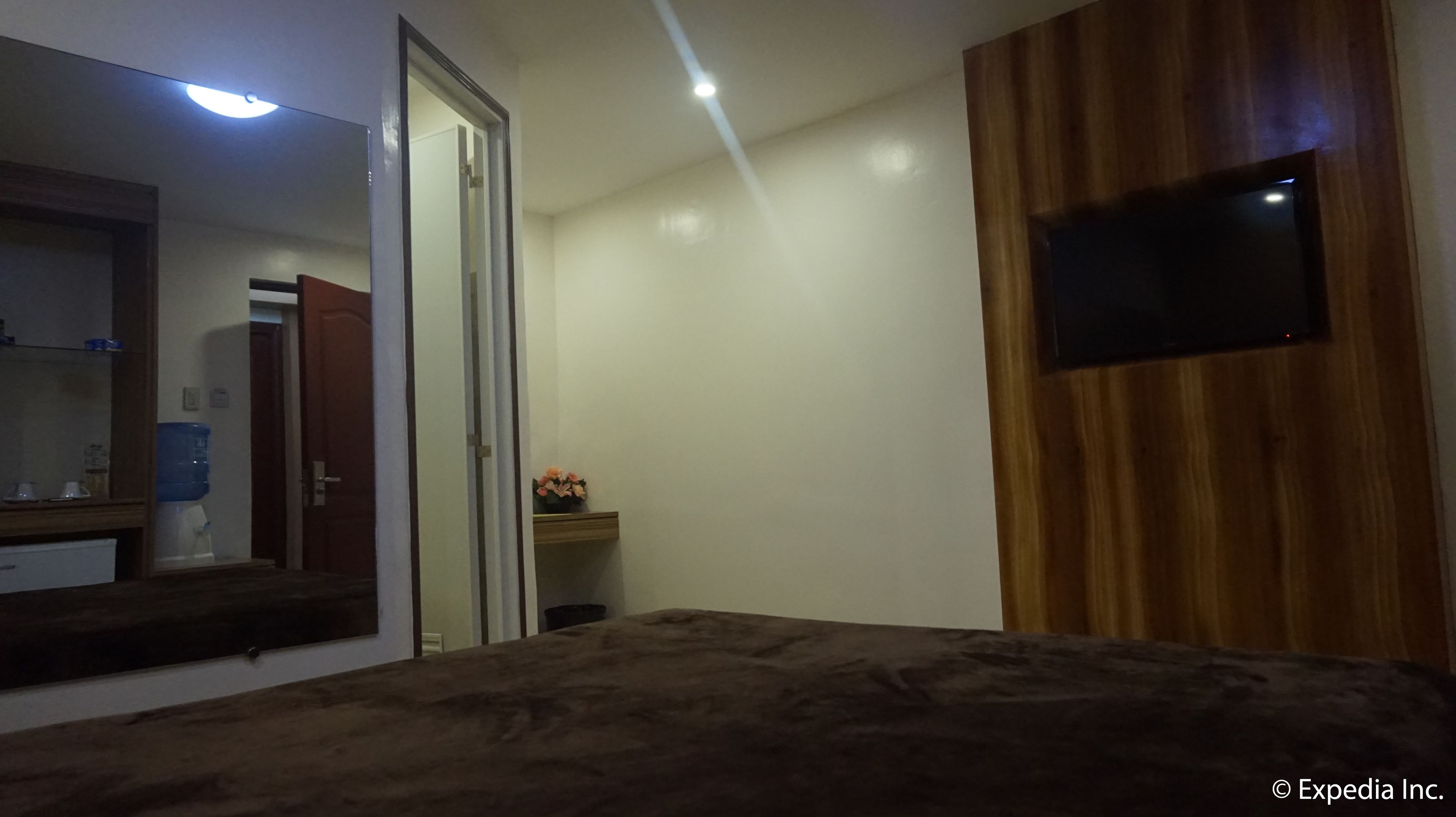Um quarto com uma cama, televisão na parede, um espelho, uma porta e uma pequena mesa com um vaso de flores.