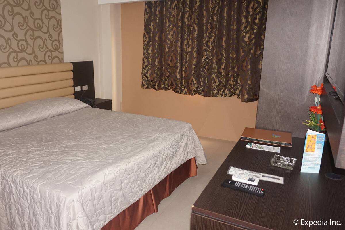 Una habitación de hotel con una cama grande, una mesita de noche de madera, un escritorio con un florero y una ventana con cortinas estampadas.
