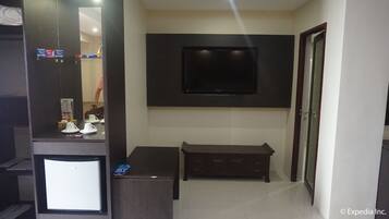 A hotel room with a flat-screen TV mounted on the wall, a small wooden bench, and a desk with a mirror and a name tag.