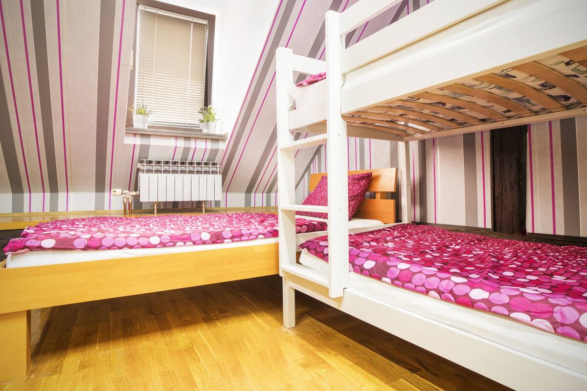 Ein Zimmer mit zwei Etagenbetten, einem Fenster mit Jalousien, einem Heizkörper und vertikal gestreiften Wänden.