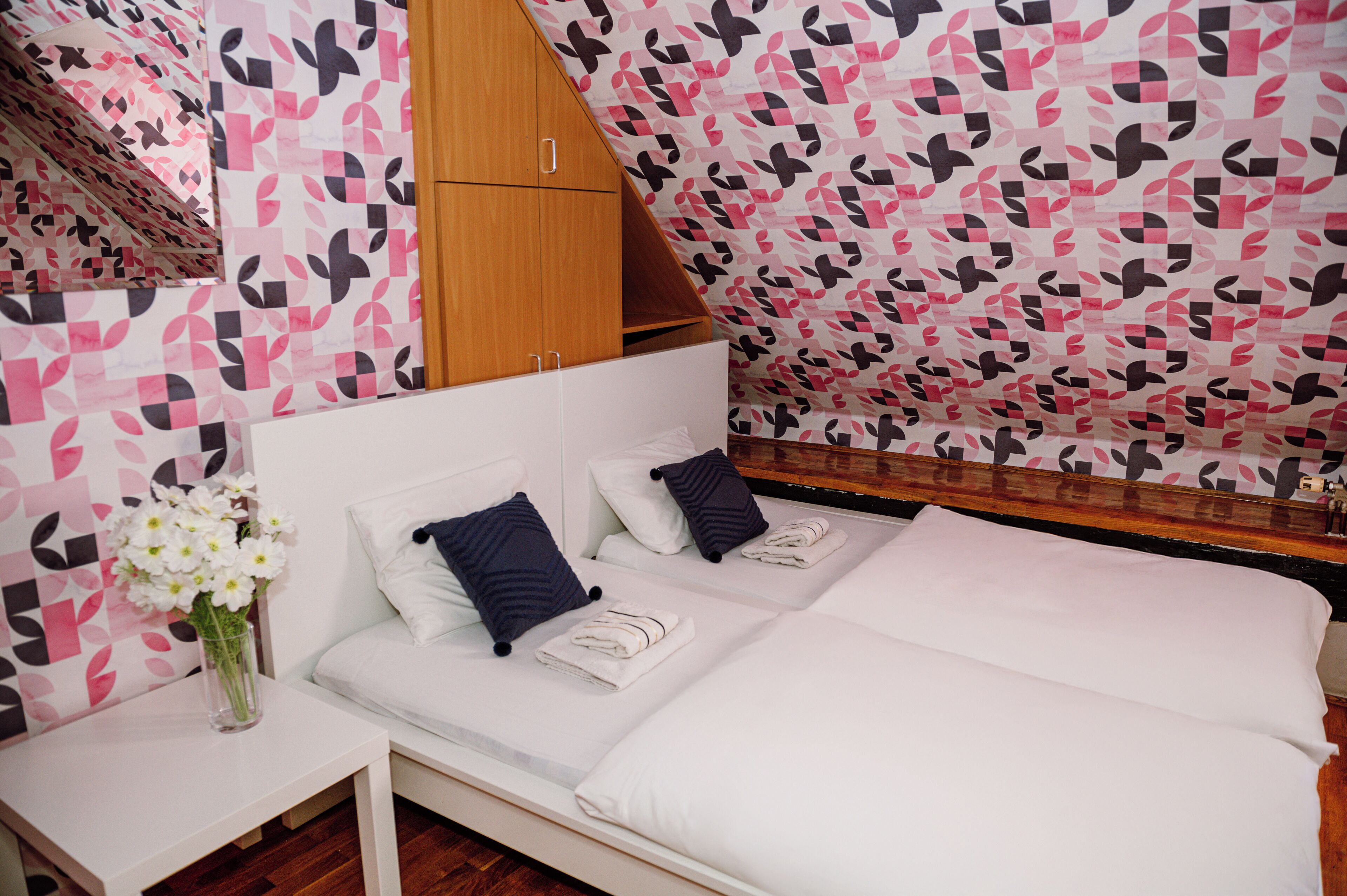 Un dormitorio con cama, dos almohadas, una mesita de noche y un jarrón con flores.