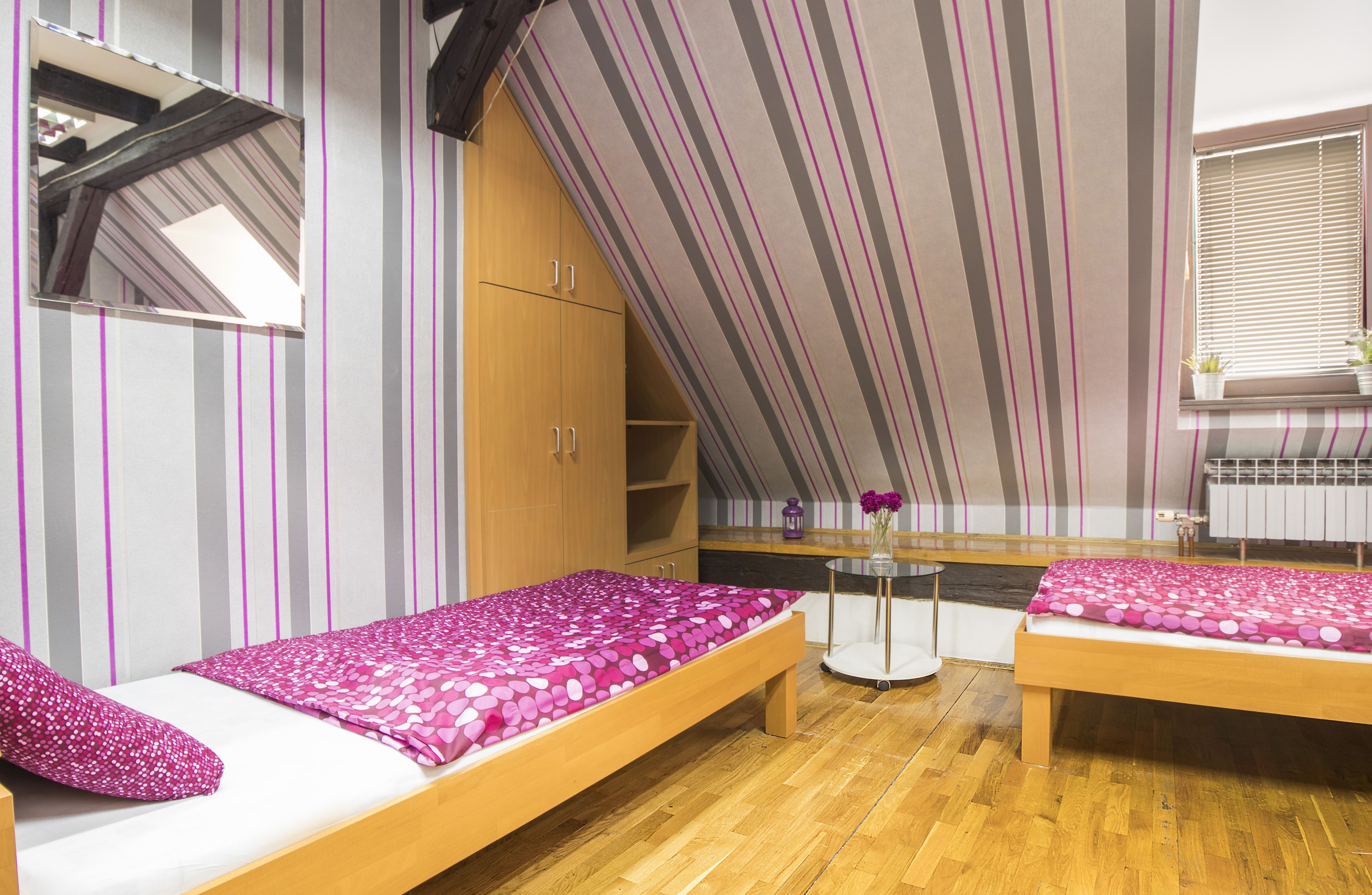 Un dormitorio con dos camas, un armario de madera, una mesita con un florero y una ventana con persianas.