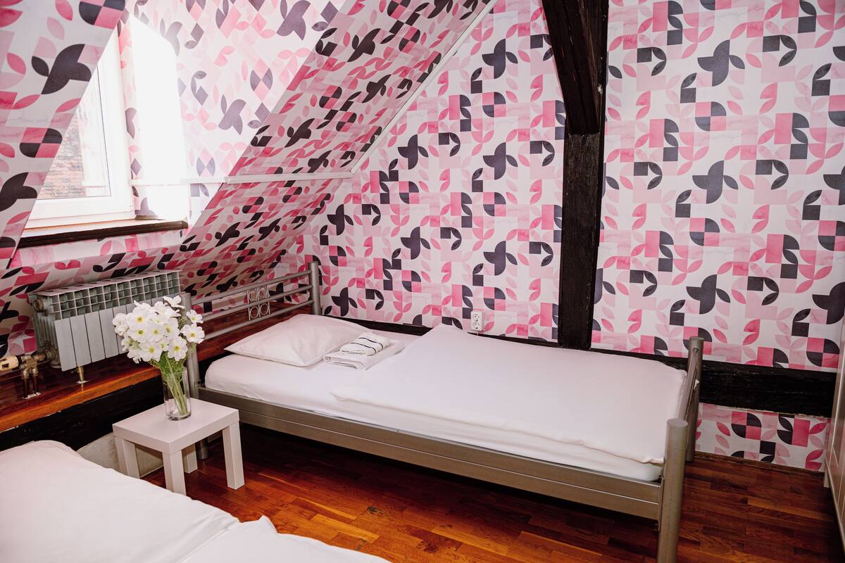 Ein Einzelbett mit weißer Matratze und Kissen, ein kleiner Nachttisch mit Blumen in einer Vase und ein Heizkörper an der Wand.