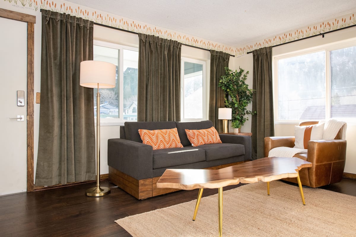 A modern living room with a sofa, a coffee table, and a chair. There are curtains on the windows, a floor lamp, and a plant.