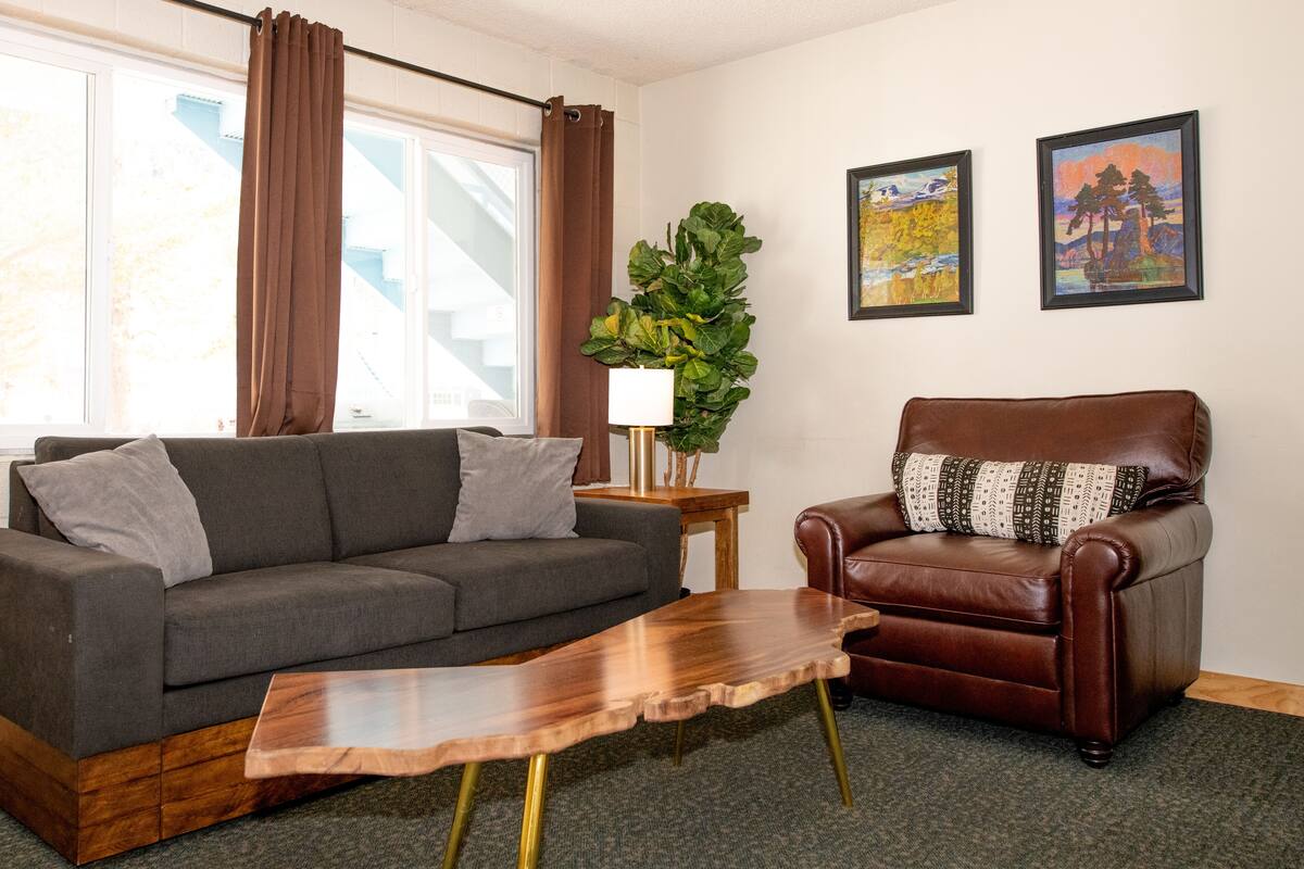 A living room with a grey sofa, a brown leather armchair, a wooden coffee table, and two framed paintings on the wall.