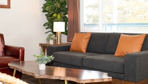 A living room with a grey sofa, two orange cushions, a wooden coffee table, a red leather chair, a small white table with a plant, and a large green plant in the background. - Gull Lake Lodge (June Lake)
