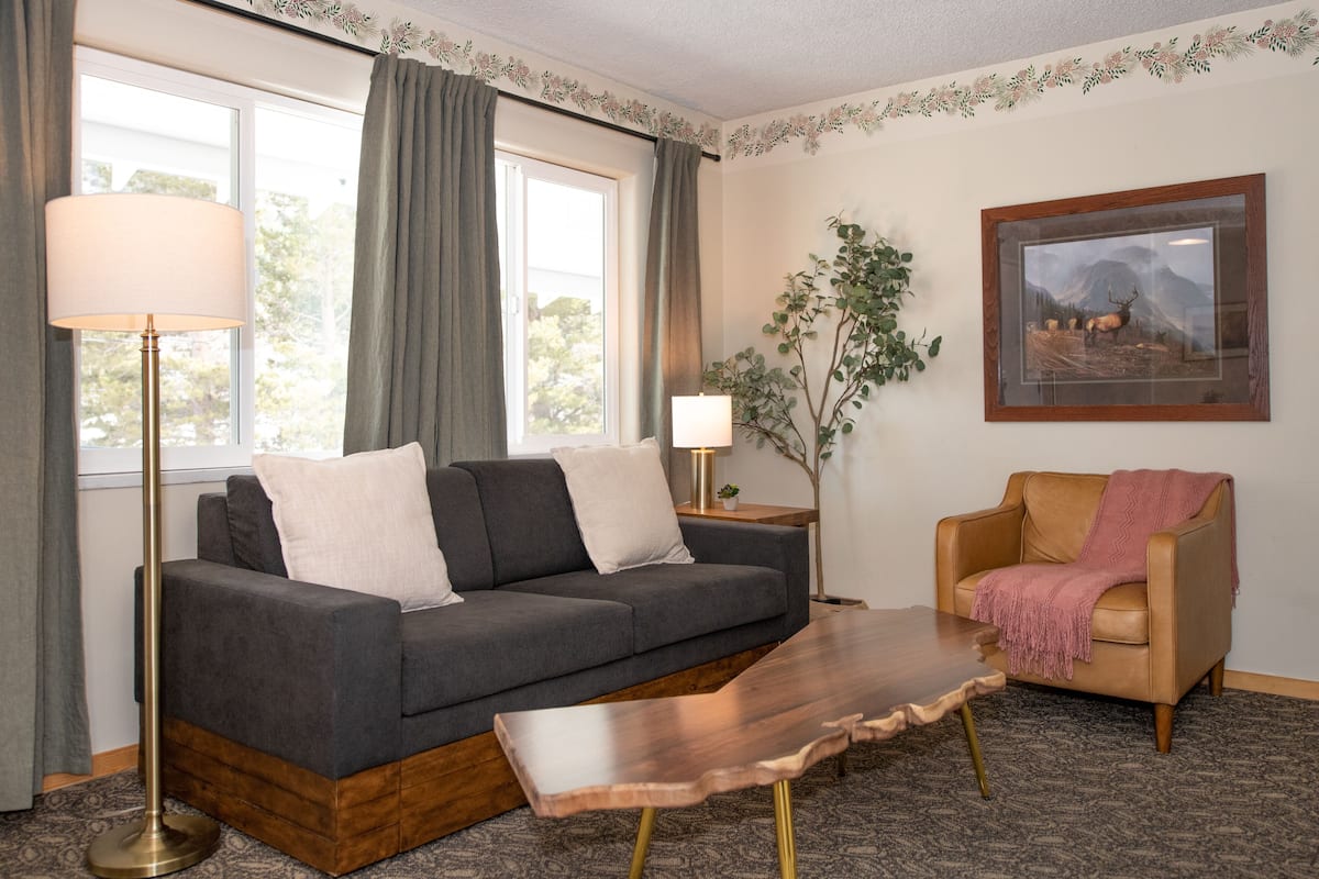 A living room with a grey sofa, a wooden coffee table, a floor lamp, a side table with a lamp, a framed picture on the wall, and a plant.
