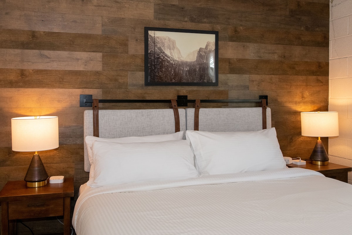 A hotel room with a wooden headboard, two bedside lamps, a framed picture on the wall, and a neatly made bed with white linens.