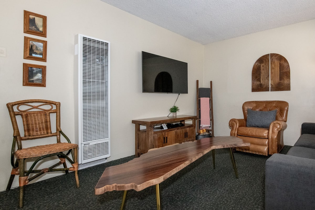 A living room with a flat-screen TV, a leather armchair, a sofa, a wooden coffee table, and a potted plant.
