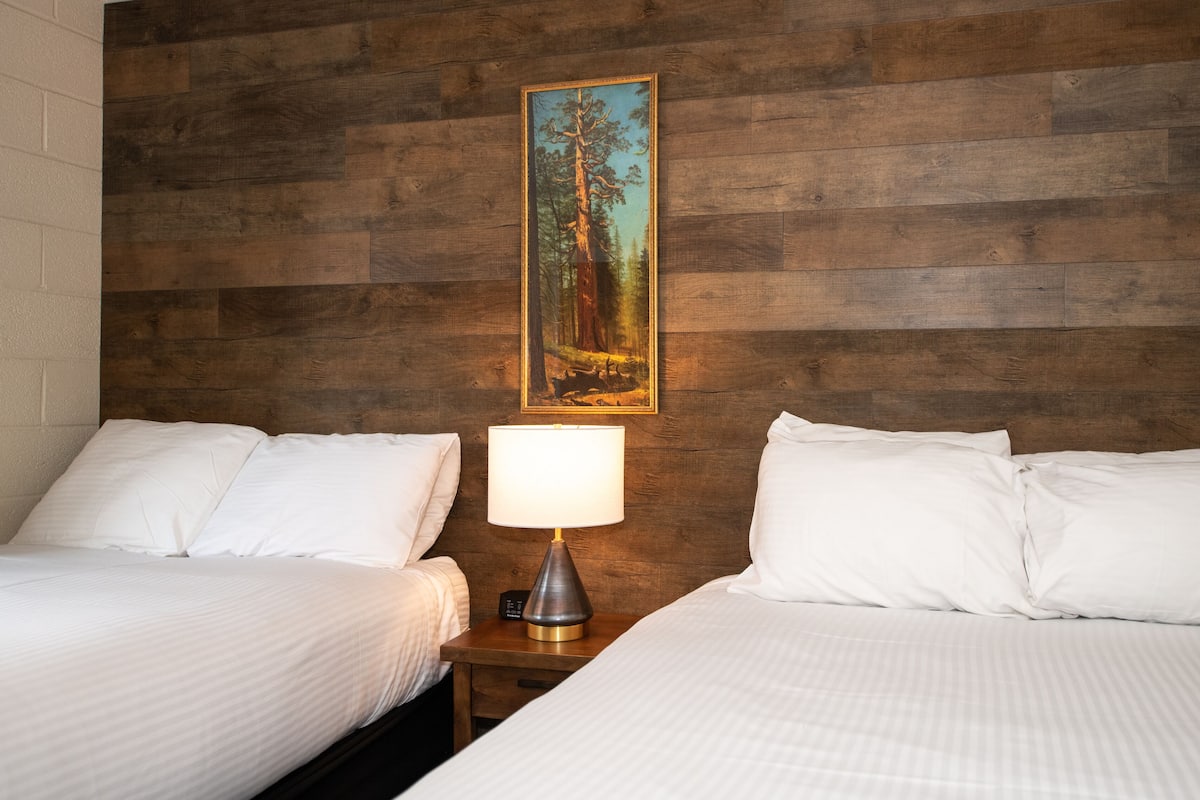 A hotel room with two beds, a nightstand with a lamp, and a painting of a forest on the wooden wall.