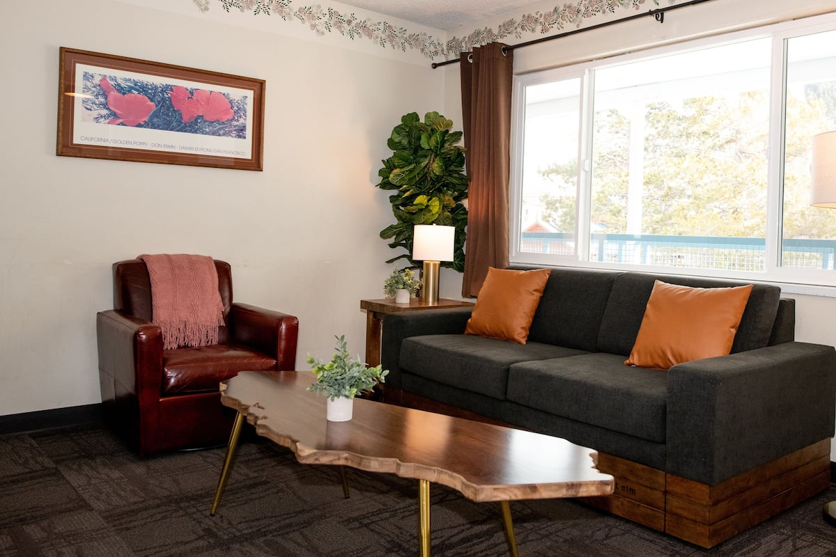 A living room with a leather armchair, a grey sofa with orange cushions, a wooden coffee table, a floor lamp, and a large window with curtains.