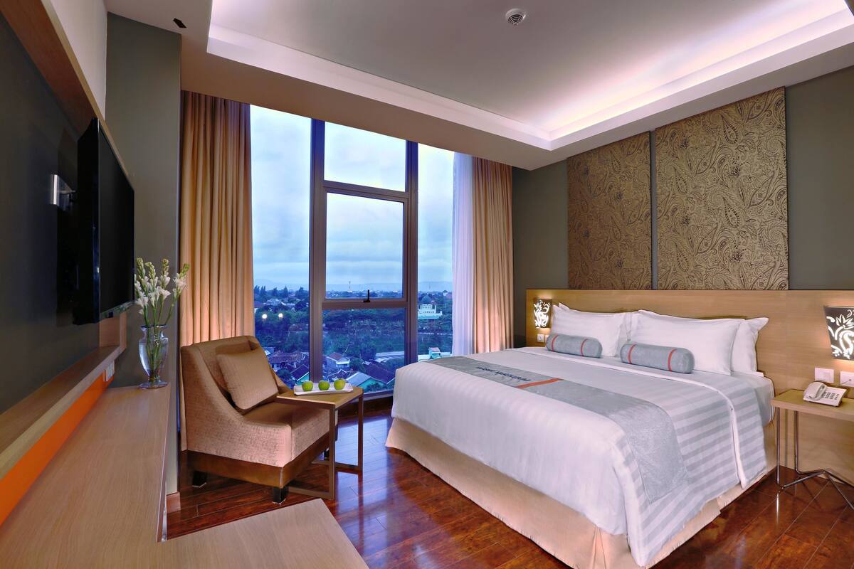 Habitación de hotel moderna con una cama grande, un escritorio, una silla y vistas a la ciudad a través de amplias ventanas.