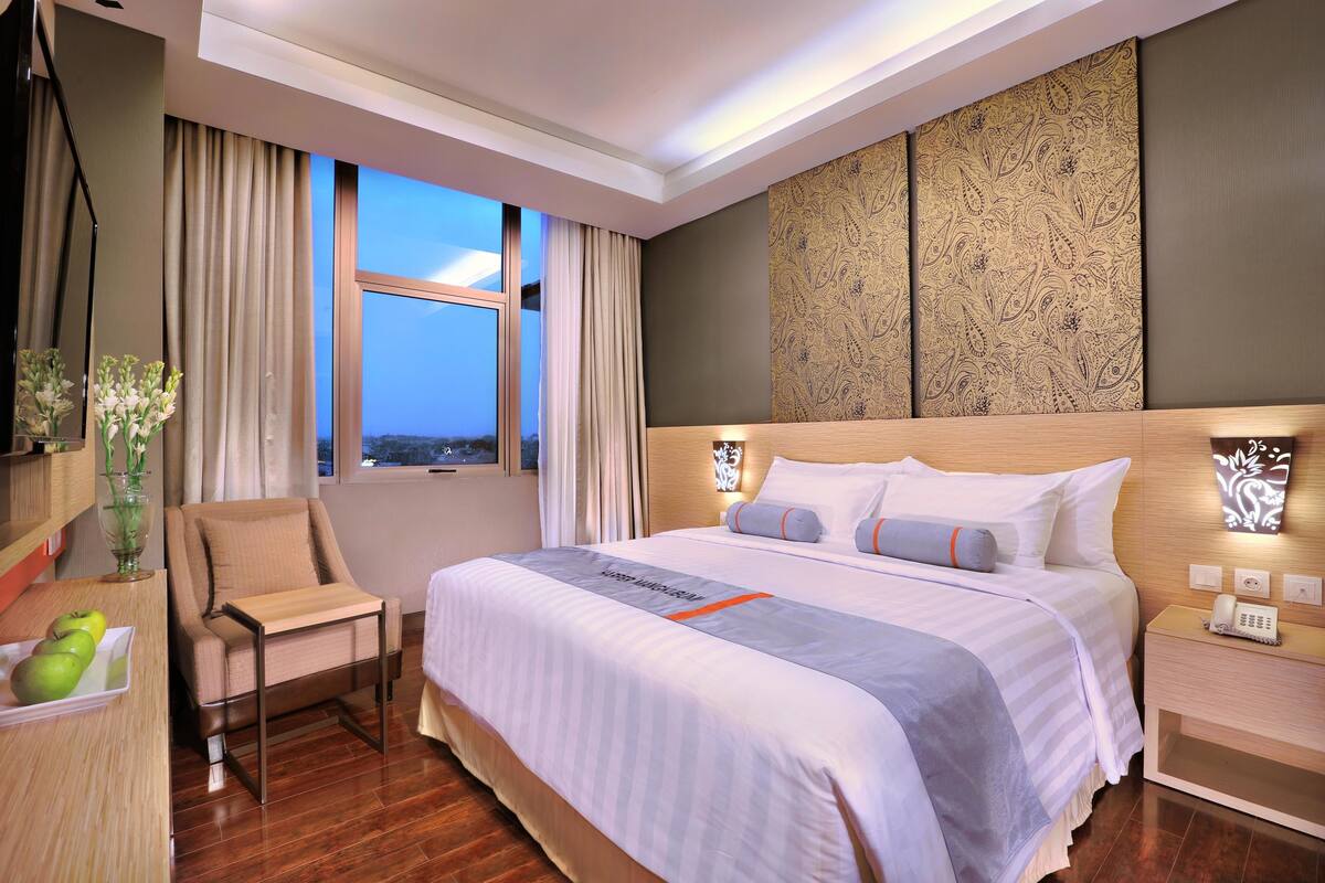 Habitación de hotel con una cama grande, una silla, una mesita de noche con lámpara y vistas a la ciudad por la ventana.