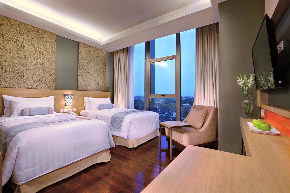 Habitación de hotel con dos camas, un escritorio, una silla, un televisor y un ventanal con vista a la ciudad.