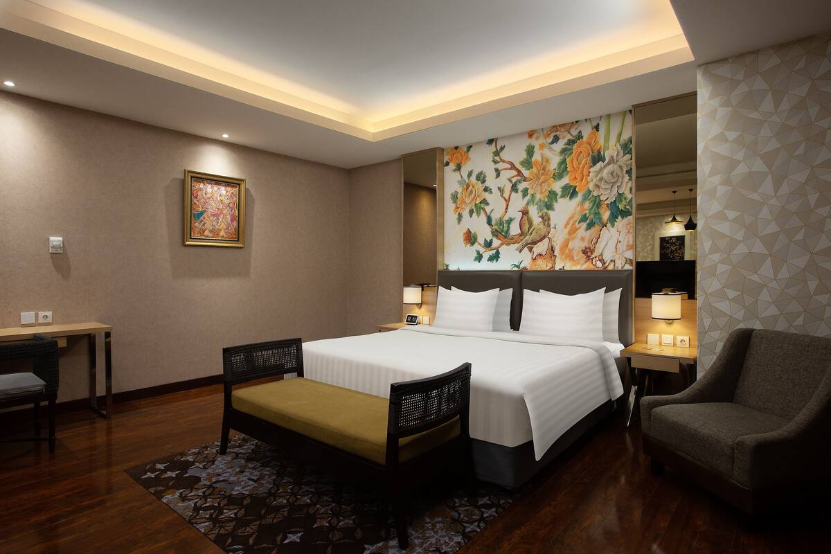 Una habitación de hotel con una cama grande, un escritorio con una silla, un banco, otra silla y una mesita de noche con lámpara.