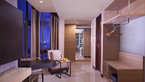 Una habitación de hotel moderna con un ventanal grande, una zona de estar con una mesa y sillas, un banco de madera y un armario con perchero.
