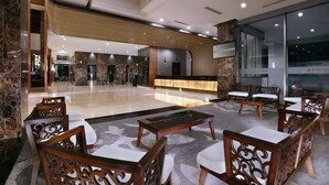 Un vestíbulo amplio con mostrador de recepción, área de estar con sillas y mesas de madera, y un diseño moderno con pisos de mármol y detalles en madera.