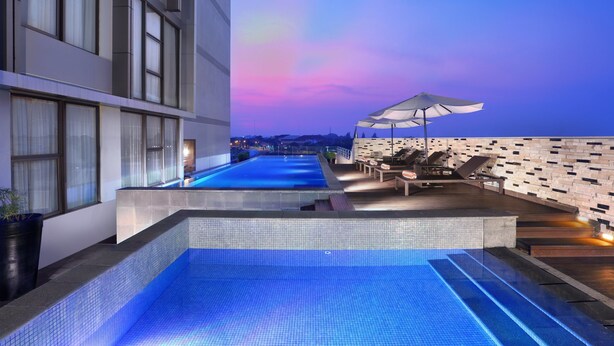 Una piscina infinita en la azotea con vistas al horizonte de la ciudad al atardecer, rodeada de sillones de descanso y una fachada de edificio moderna.