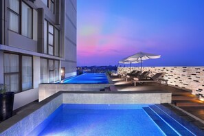 Una piscina infinita en la azotea con vistas al horizonte de la ciudad al atardecer, rodeada de sillones de descanso y una fachada de edificio moderna.