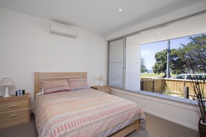 A bedroom with a large window, a bed with patterned bedding, bedside tables with lamps, and a view of the outdoors. - Two Bays Apartments (Torquay)