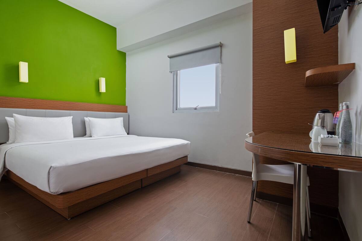 Ein Hotelzimmer mit Bett, einem kleinen Tisch, einem Fenster mit Jalousie und einer grünen Wand.