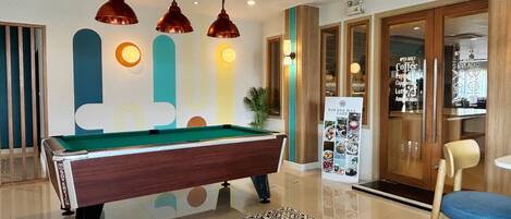 A pool table in a well-lit room with pendant lighting, a patterned rug, and a door leading to a cafe.