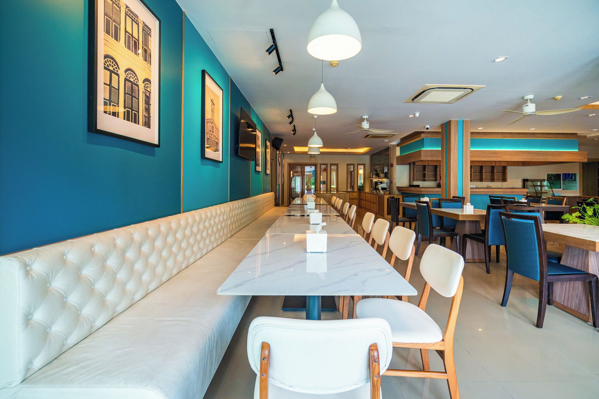 A long dining table with white cushioned benches, framed pictures on teal walls, and modern pendant lighting.