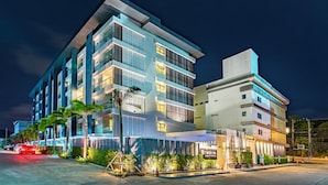 A multi-story hotel with a modern design, featuring balconies, a well-lit entrance, and a landscaped area with palm trees. - Ratana Hotel Rassada (Ratsada)
