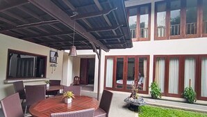 A covered outdoor dining area with a round table and wicker chairs, a small garden with a fountain, and a poolside view. - Sekuta Condo Suites (Denpasar)