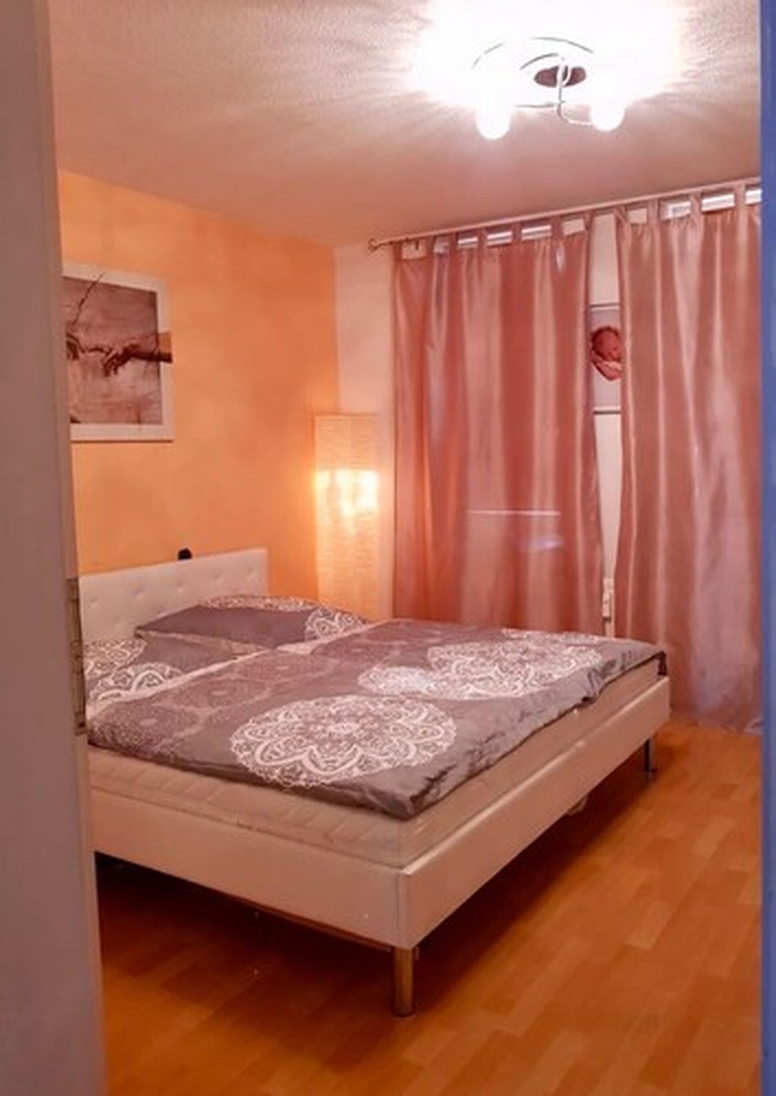 A bedroom with a bed, curtains, a wall-mounted lamp, and a ceiling light fixture.