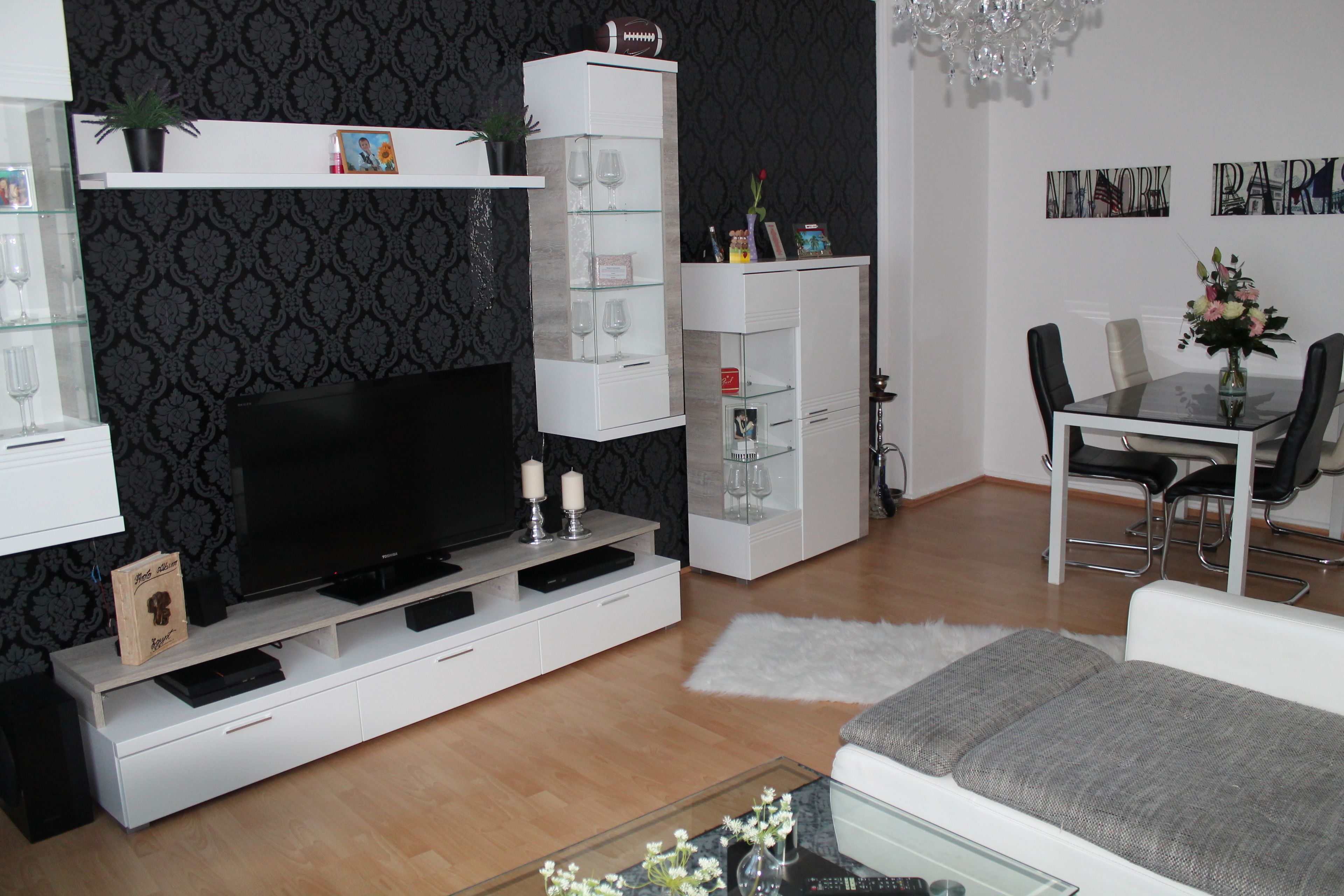 Una sala de estar moderna con televisor de pantalla plana, mueble blanco para equipo electrónico, estanterías de vidrio y un comedor con mesa y sillas.