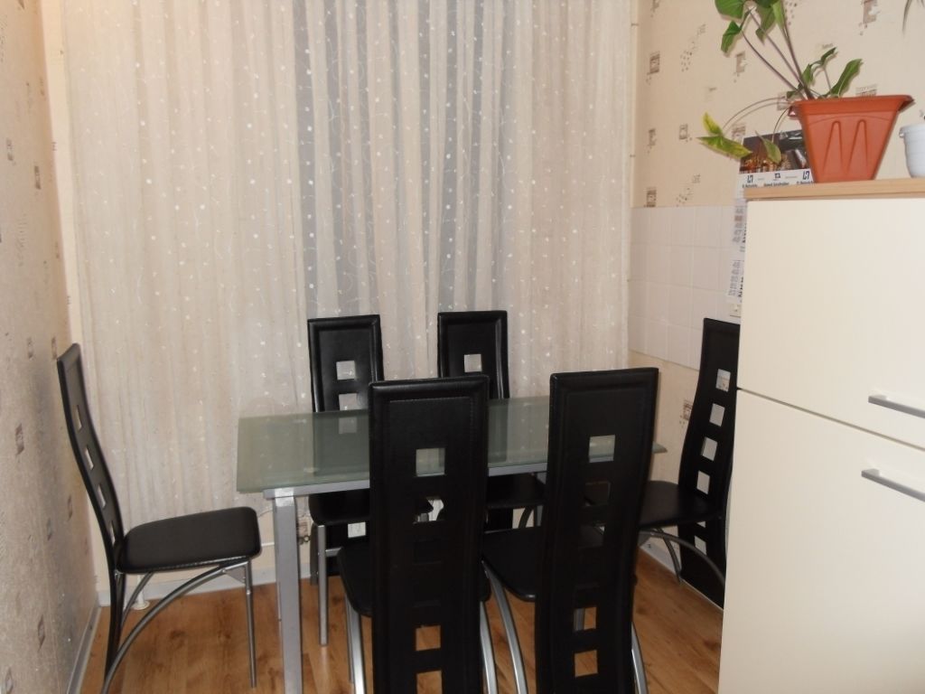 Un pequeño comedor con una mesa de vidrio y cuatro sillas negras. A la derecha, un armario blanco con dos cajones.