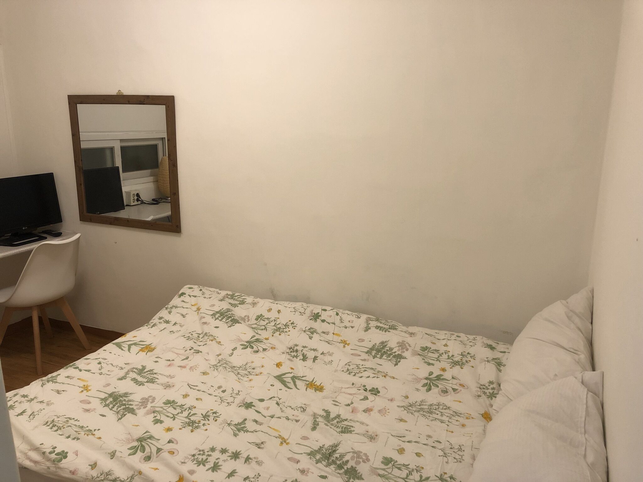 A small, sparsely furnished room with a single bed, a desk with a computer, and a mirror on the wall.