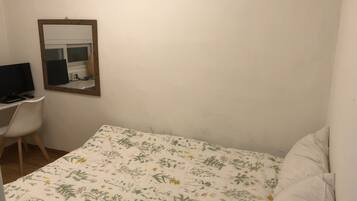 A small, sparsely furnished room with a single bed, a desk with a computer, and a mirror on the wall.