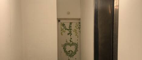 Un pasillo estrecho con una pared blanca, una puerta adornada con un arreglo de hojas verdes y un corazón con el texto "Love in the world, Love on earth", y una puerta de ascensor negra.
