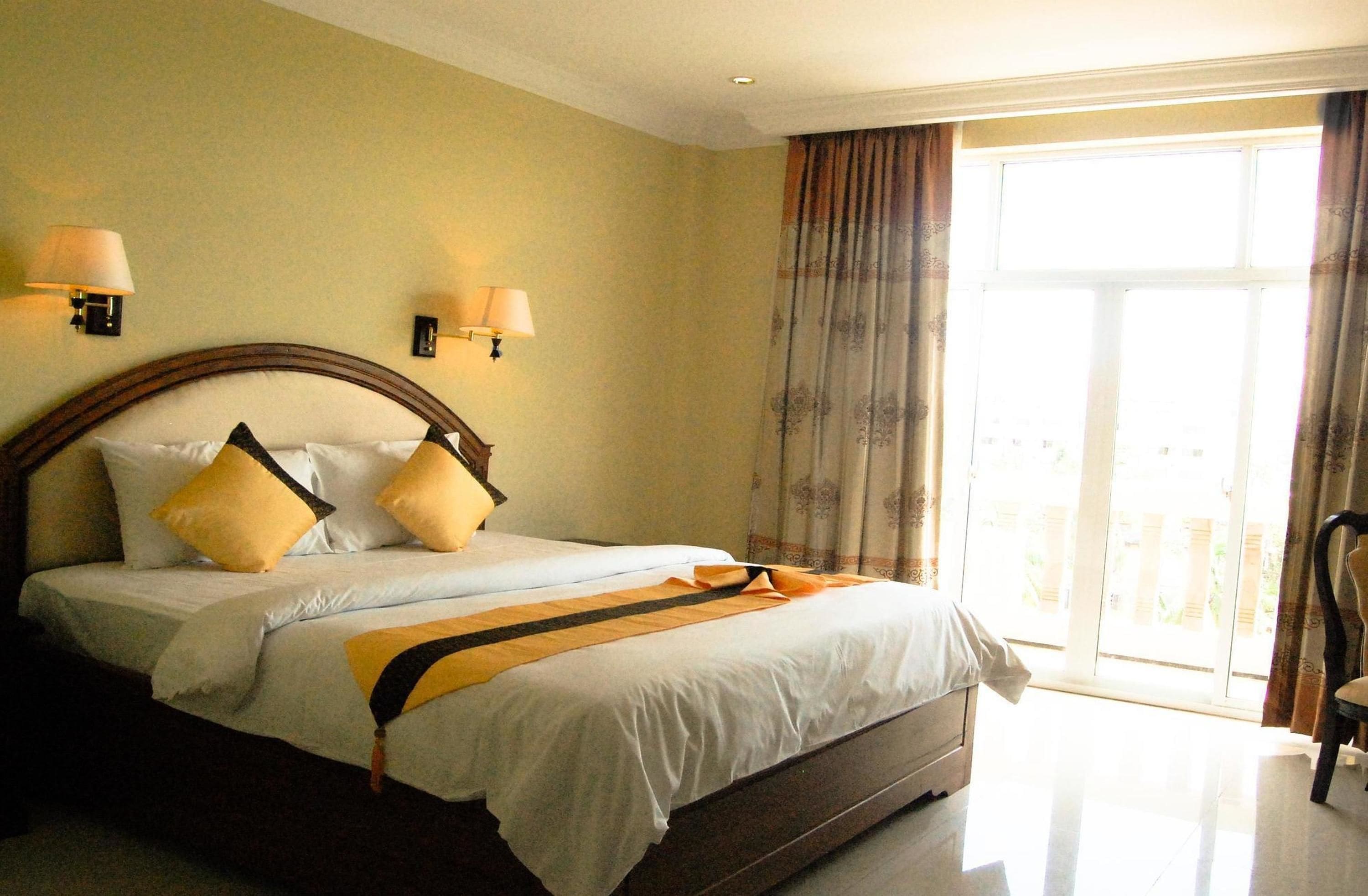 A hotel room with a large bed, two wall-mounted lamps, a window with curtains, and a chair.