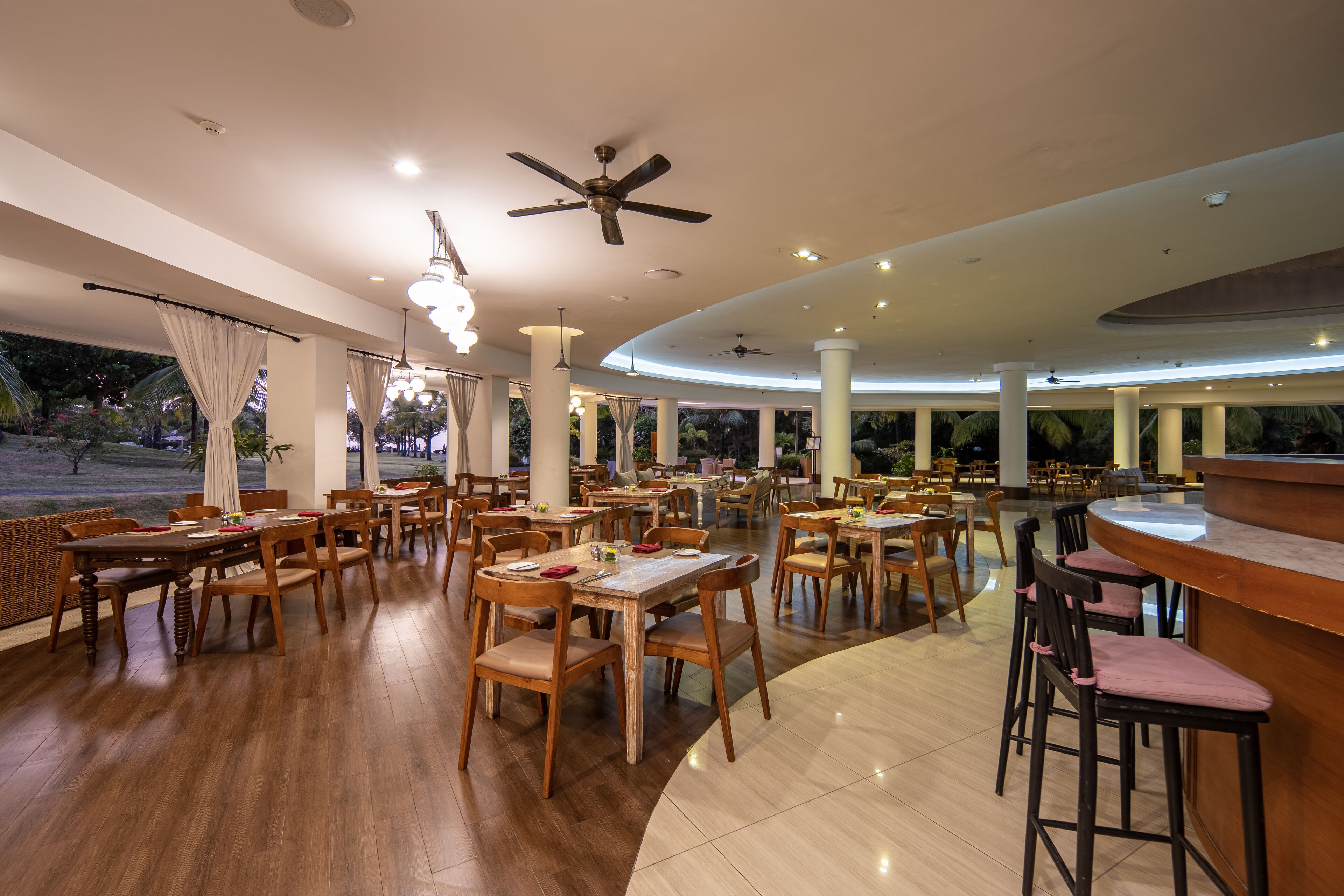 Ein großzügiges Restaurant mit Holztischen und -stühlen, Deckenventilatoren und großen Fenstern mit Blick ins Freie.