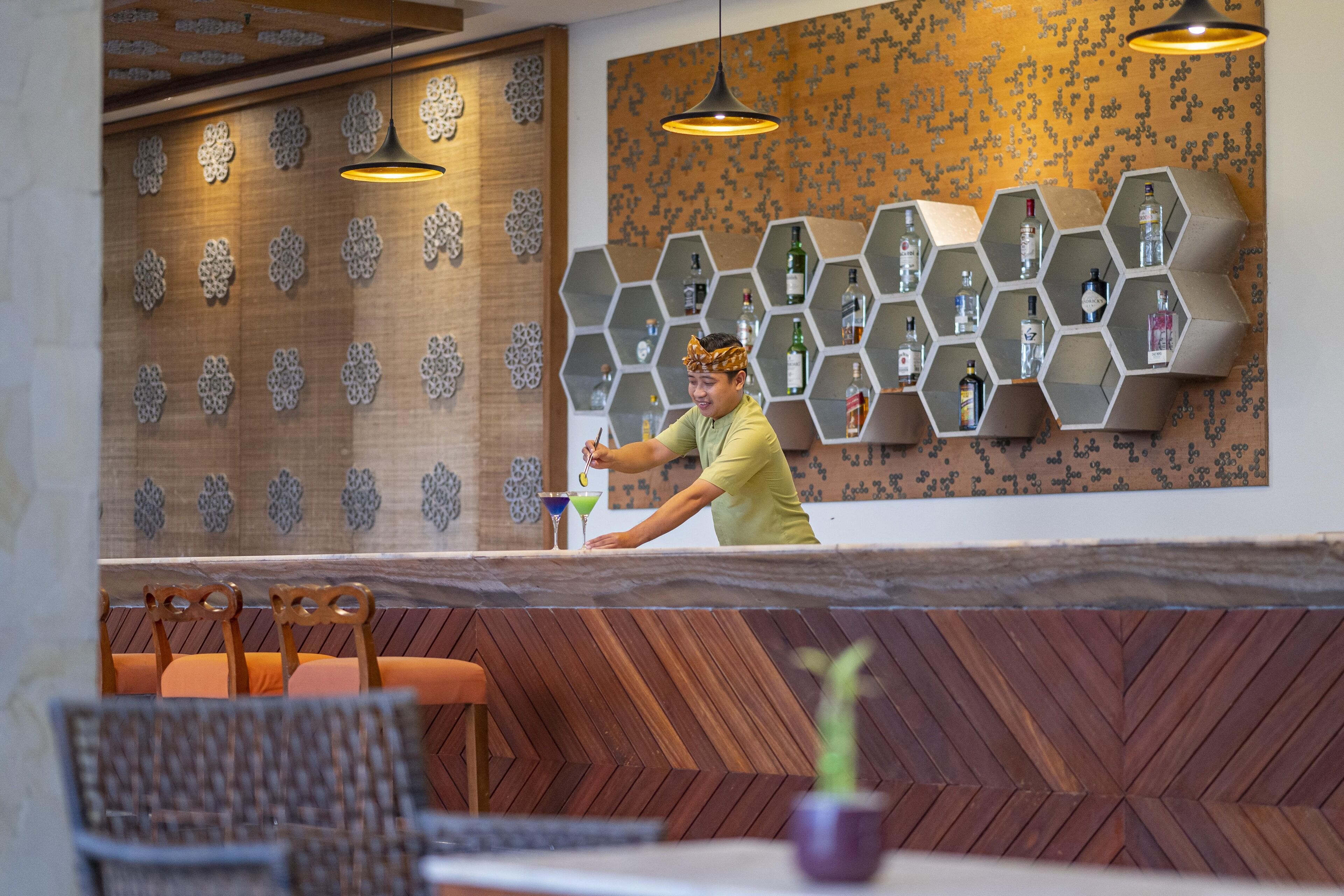 A bar area with hexagonal shelves displaying bottles, wooden wall decor, and a person preparing a drink.