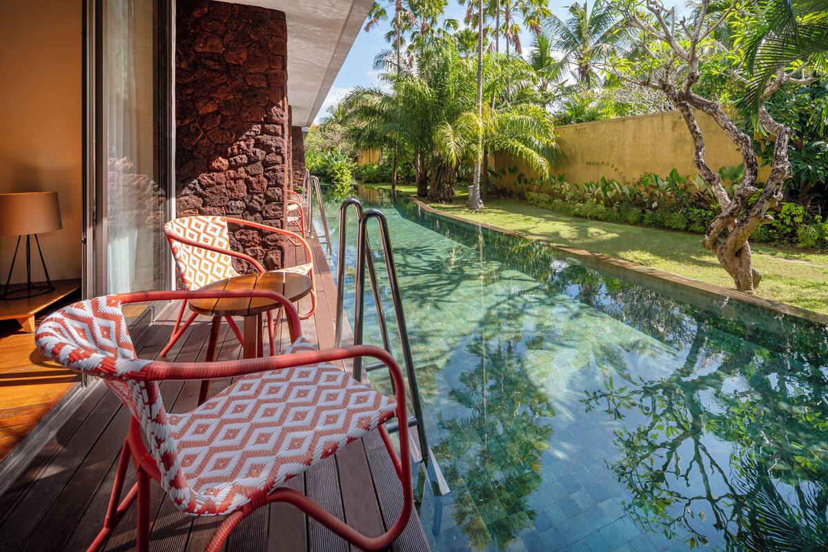 Un área junto a la piscina con una terraza de madera, una silla con estampado rojo y blanco, y una pared de piedra.
